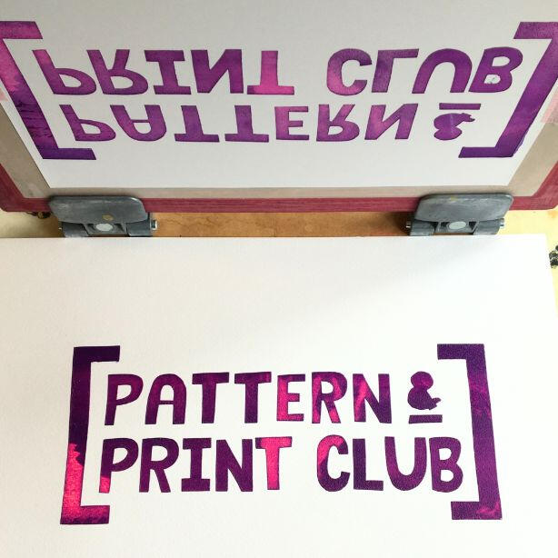 Screen printing frame held in hinges with stencil on it. Mono print image in purple and pink text saying Pattern & Print Club with brackets on each end of the text. Wooden board background.